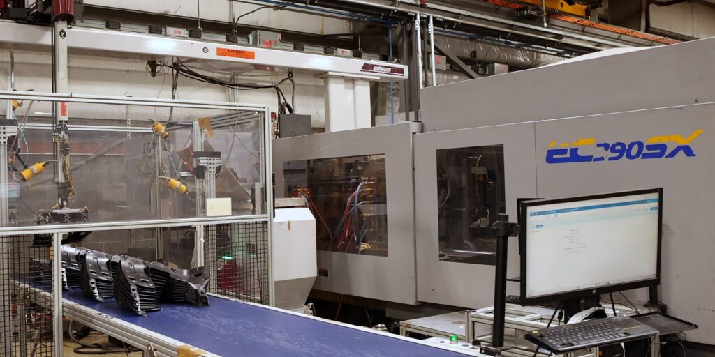 Robotic molding cell sending black molded parts down a conveyor beside an all-electric press